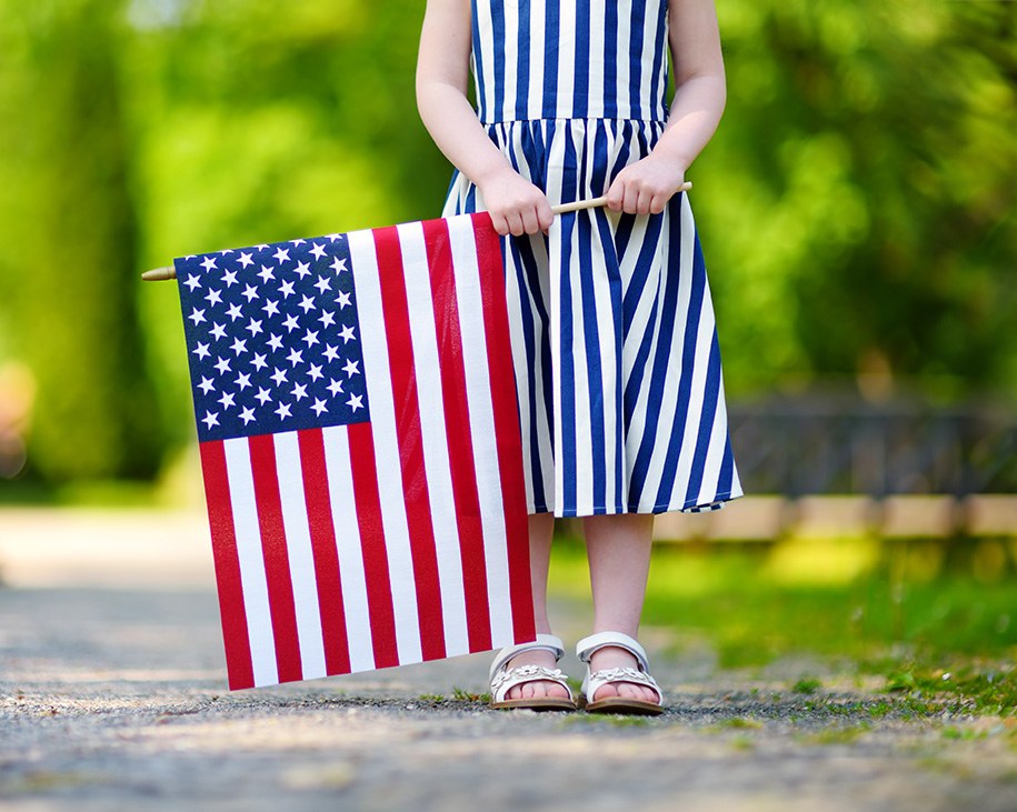 A little girl wearing a white and blue striped dress and white sandals holding a United States flag.