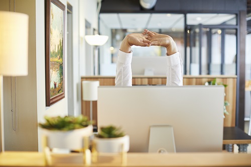 A person sitting behind a computer screen and stretching their arms over their head.