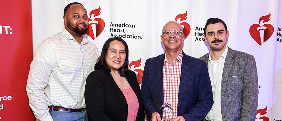 Three men and a woman pose with an award in front of a red and white backdrop with the American Heart Association logo.