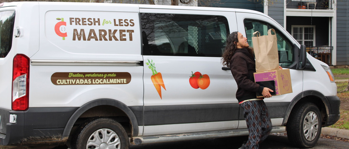 A man unloads a box and paper bag filled with food items from a van painted with the words "Fresh for Less Market" and "Cultivadas Localmente" outside a multi-unit home. 