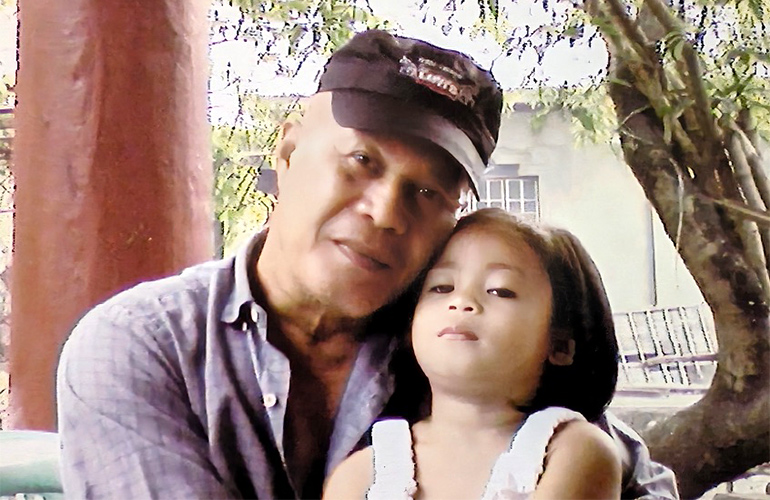 A man in a baseball cap poses with his young daughter outside under a tree